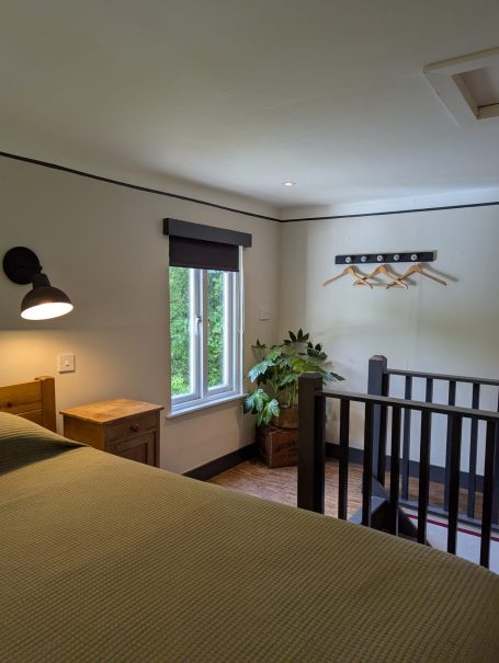 Cosy bedroom with coat rack A well-lit room with a bed, window, and wooden furnishings, featuring a staircase and coat rack.