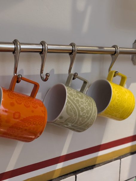 Basics provided Four ceramic mugs in orange, grey, and yellow hanging on a metal rack.