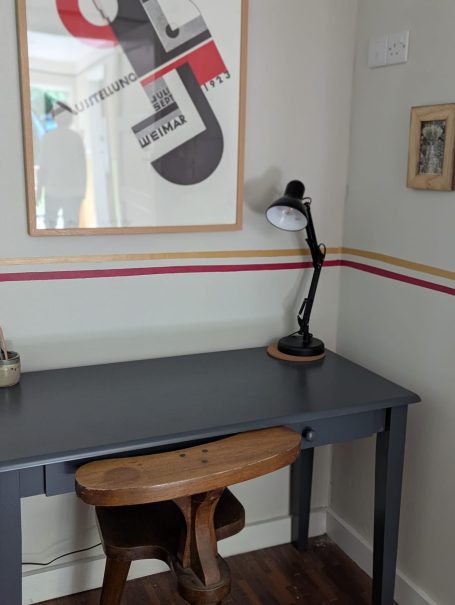 Desk A grey desk with a wooden chair, lamp, and abstract wall art in a light room.